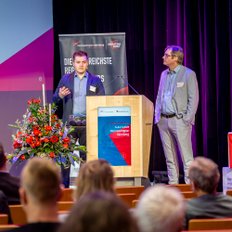 Zwei Männer halten auf der Bühne einen Vortrag.